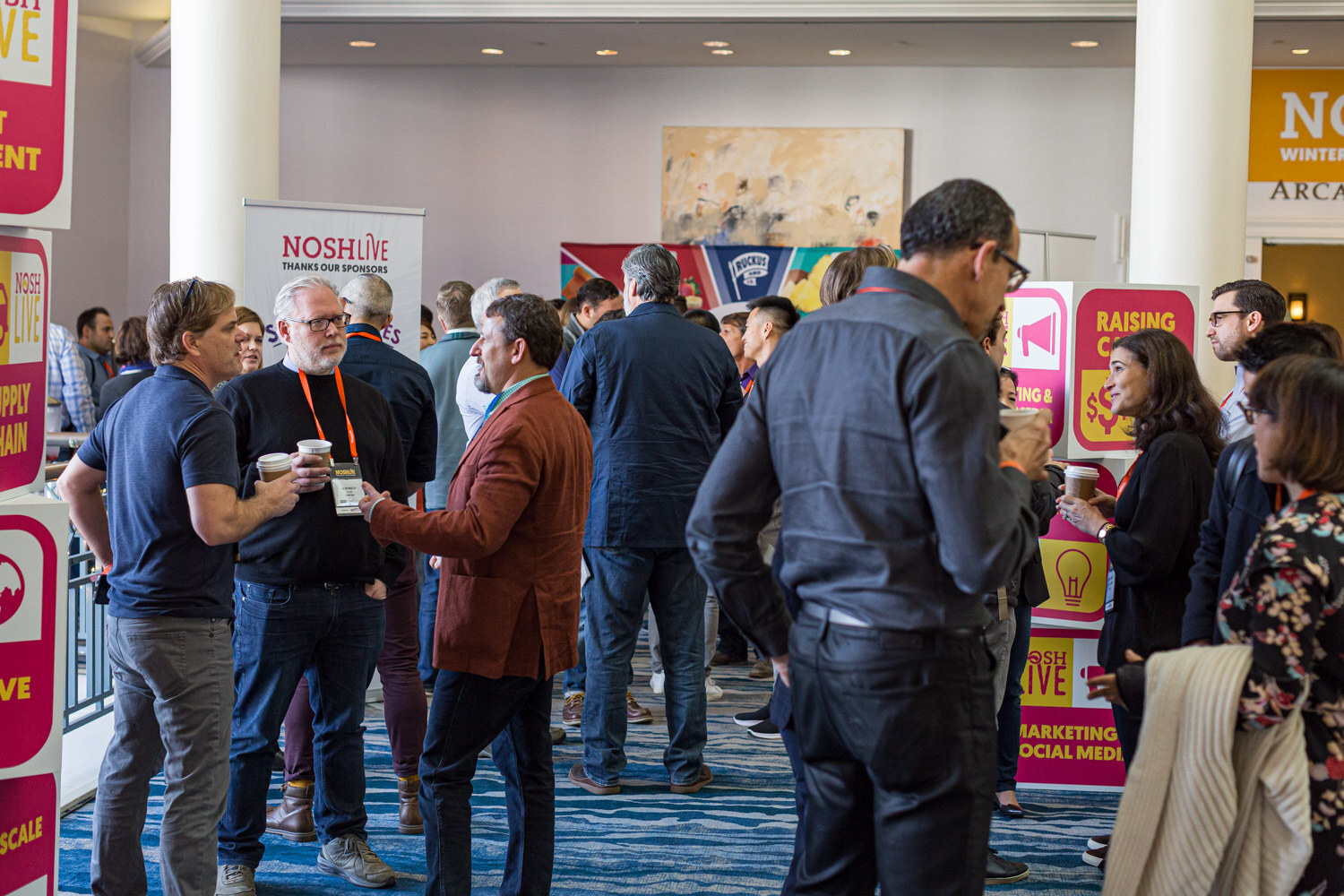 Attendees mingling with coffee in a busy foyer with Nosh Live signage