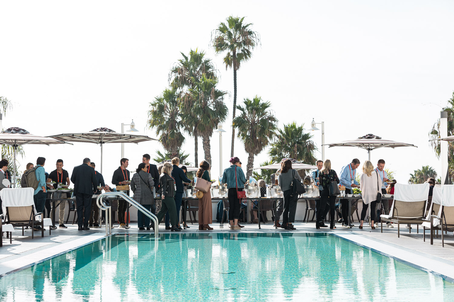 Attendees gather at lunch buffets beside a turquoise pool with palm trees and umbrellas
