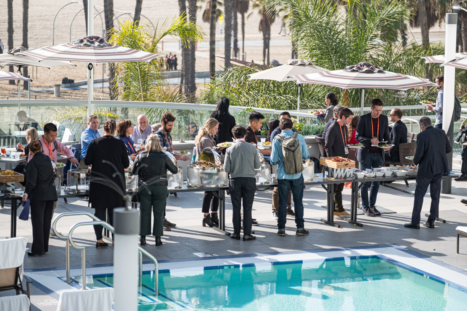 Attendees line up at buffet tables beside a swimming pool under striped umbrellas with palm trees behind