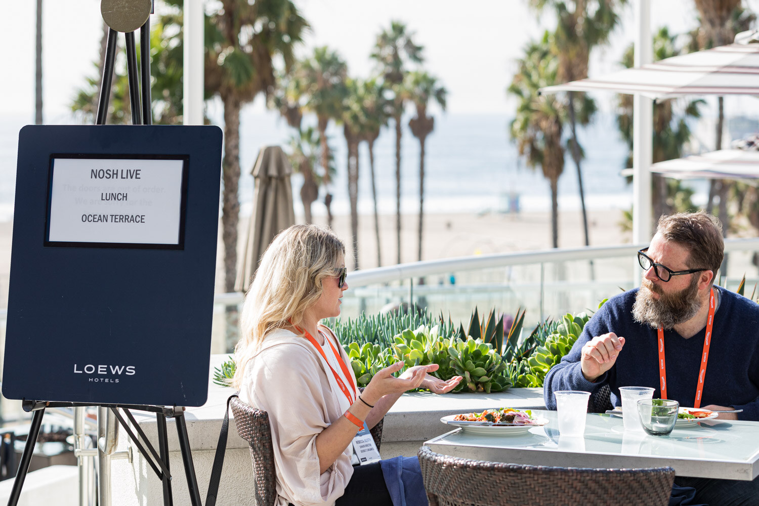 Two diners at an outdoor table near a Loews easel sign reading Nosh Live Lunch Ocean Terrace with the beach behind