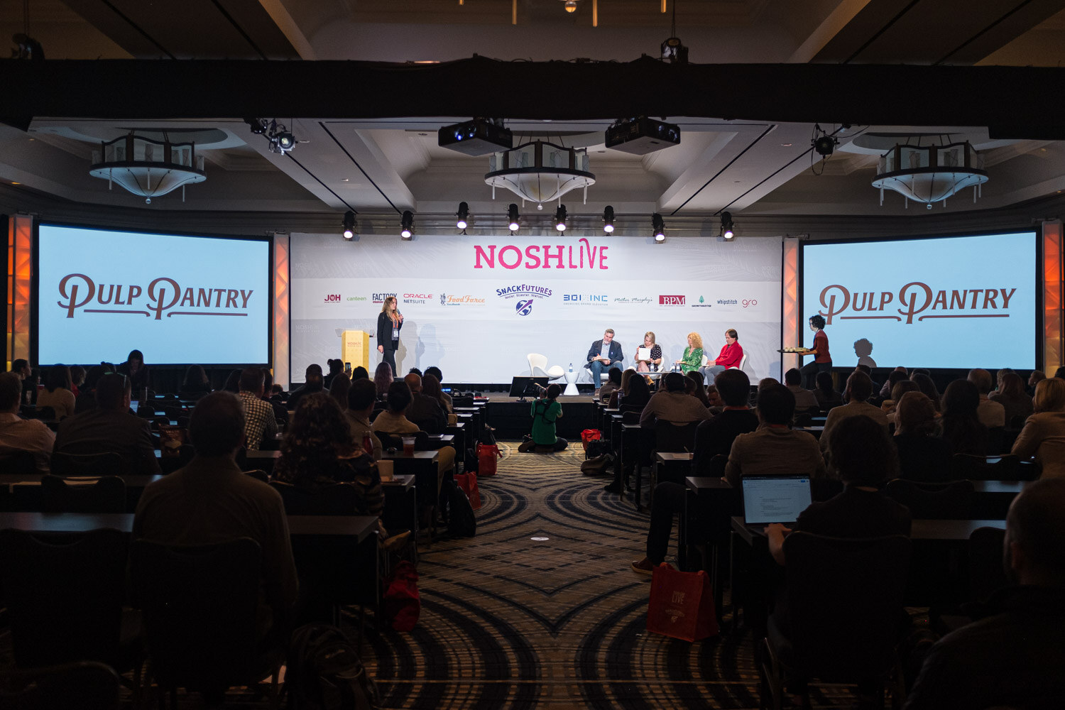 A wide view of the ballroom with Pulp Pantry logos on two side screens and the audience at tables
