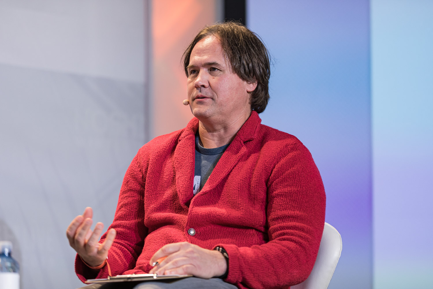 A man in a red cardigan with a headset microphone seated and gesturing with one hand