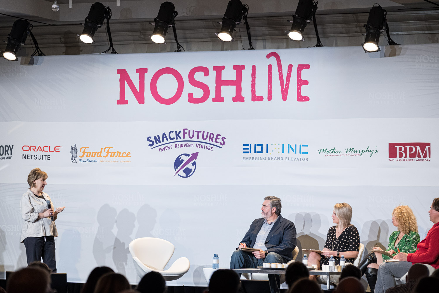 A woman standing on stage in front of four seated judges under the Nosh Live banner