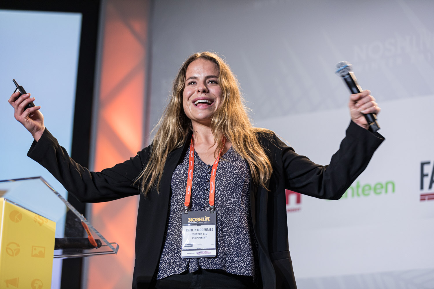 A woman in a black blazer with outstretched arms, one hand holding a microphone and the other a clicker
