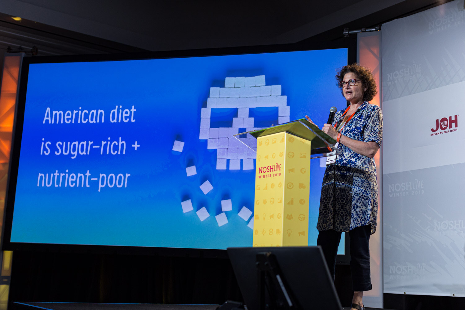 A woman at the lectern in front of a slide reading American diet is sugar plus nutrient-poor