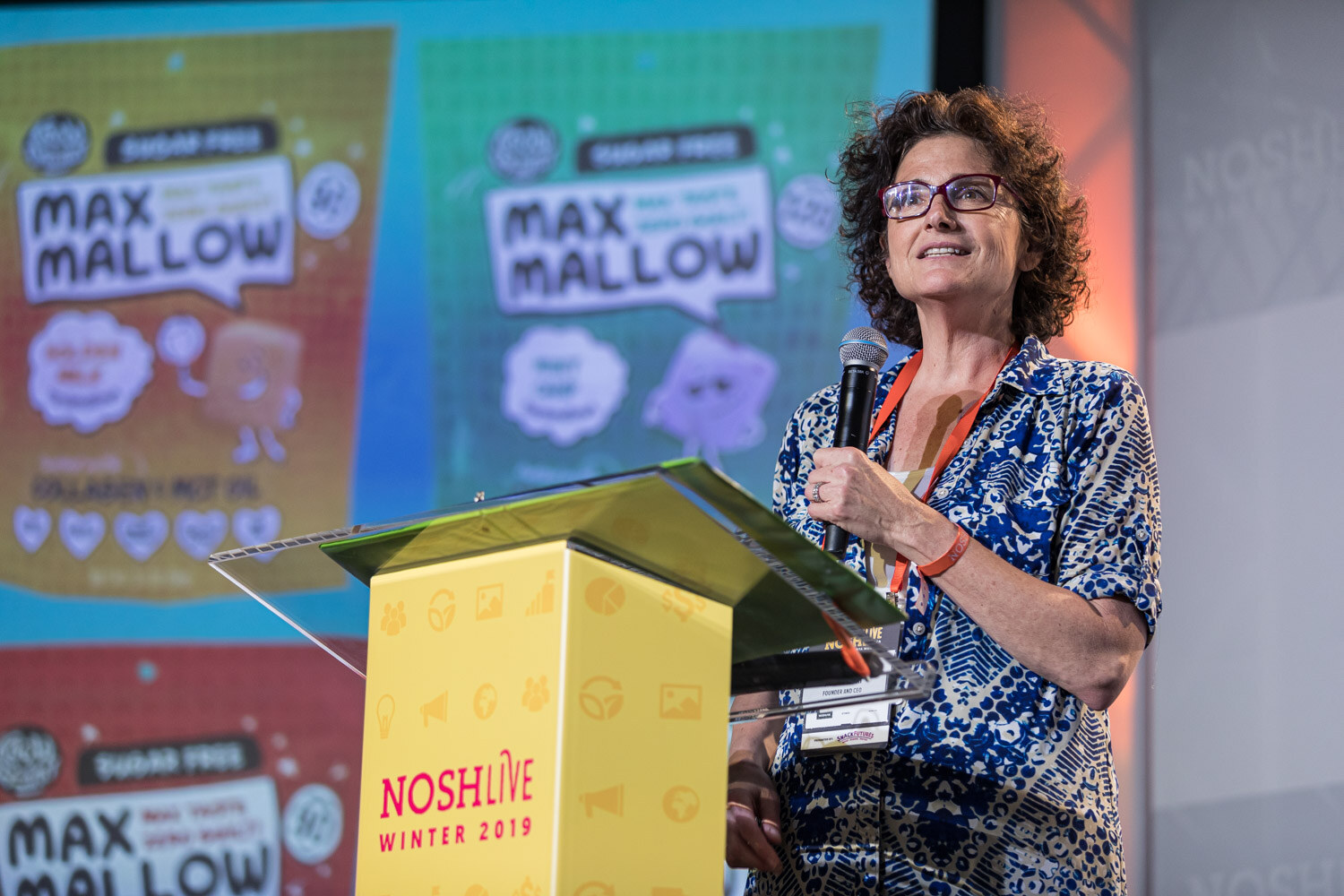 A woman with glasses at the lectern in front of a slide showing Max Mallow marshmallow packages