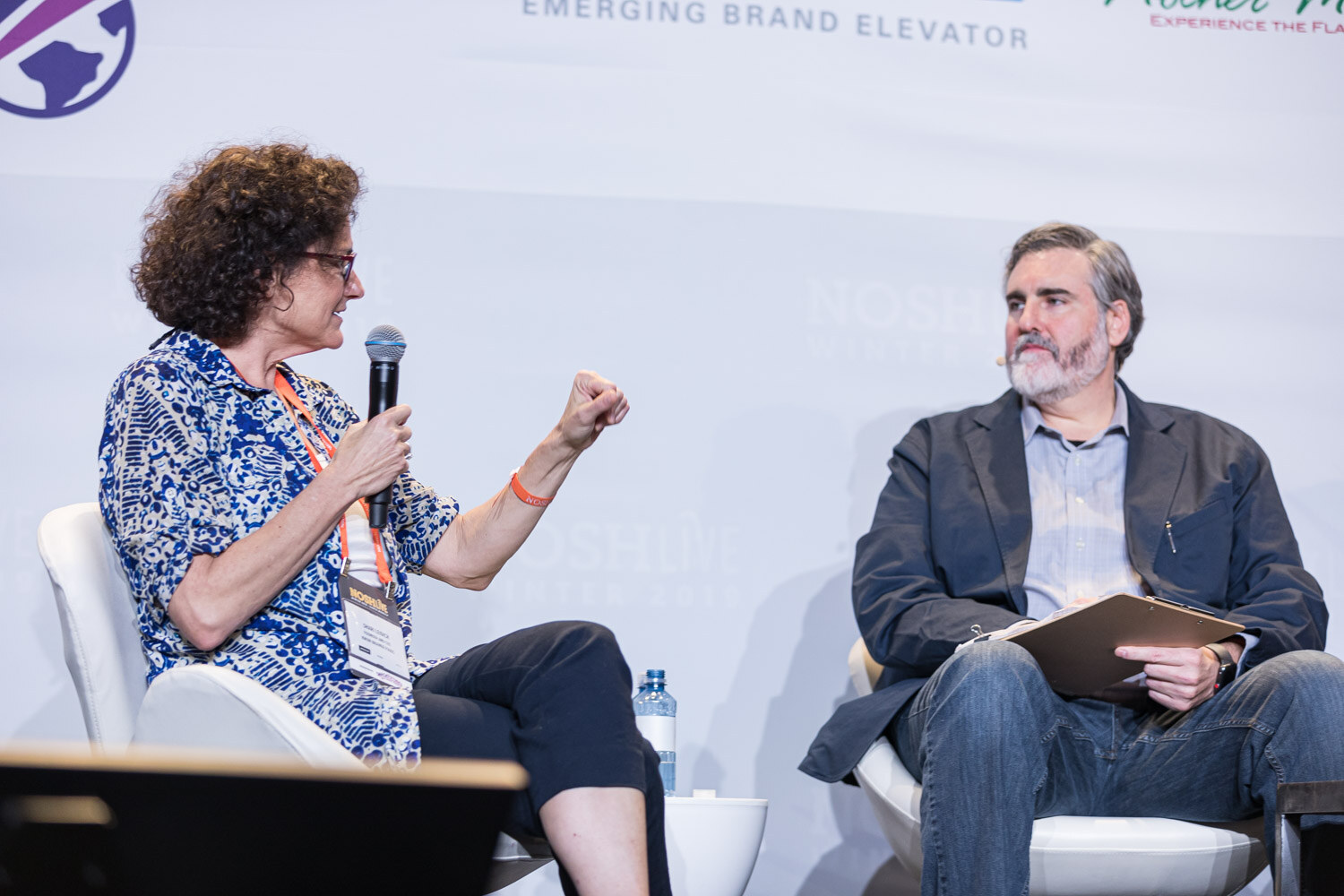 A woman in a blue patterned top speaking into a microphone and gesturing toward a bearded man holding a clipboard