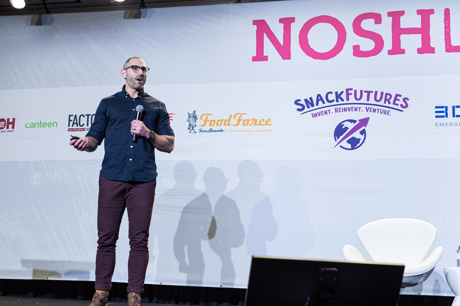 A man in a navy shirt standing at center stage in front of the Nosh Live banner