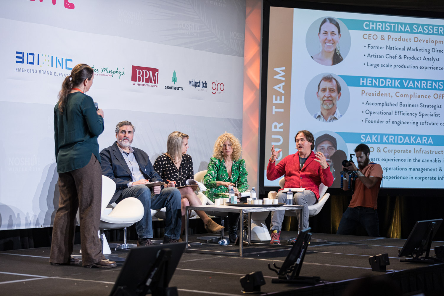 A woman standing beside four seated judges while a screen shows Our Team profiles