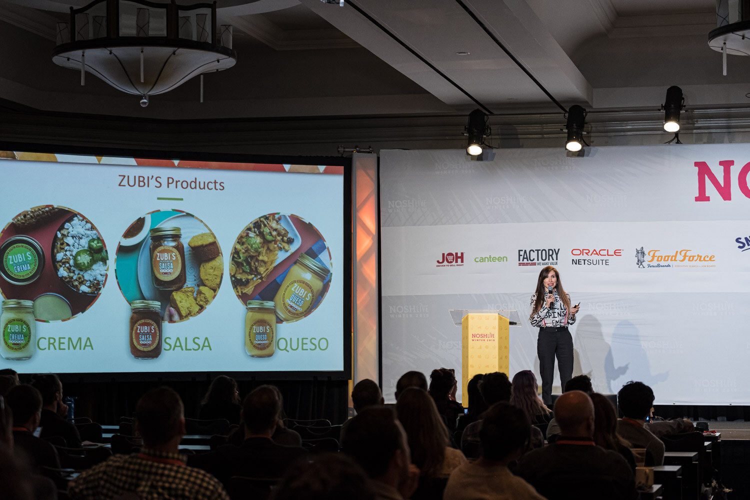 A woman at the lectern on stage with a screen displaying Zubi's Crema, Salsa and Queso jars