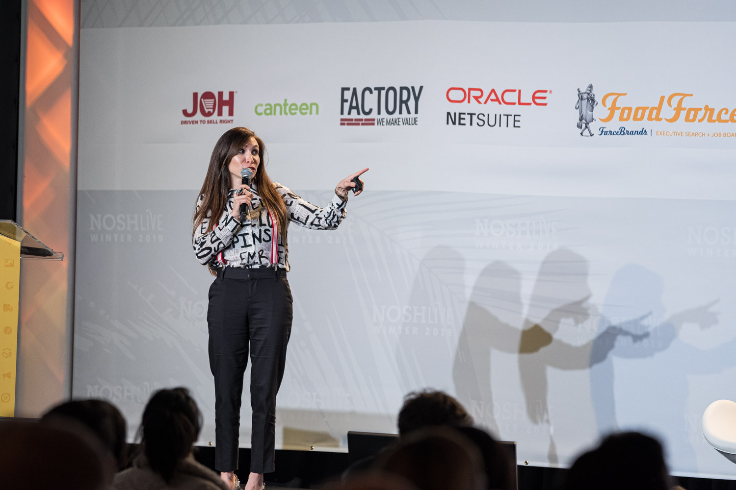 A woman in a lettered blouse and suspenders holding a microphone and pointing during her pitch