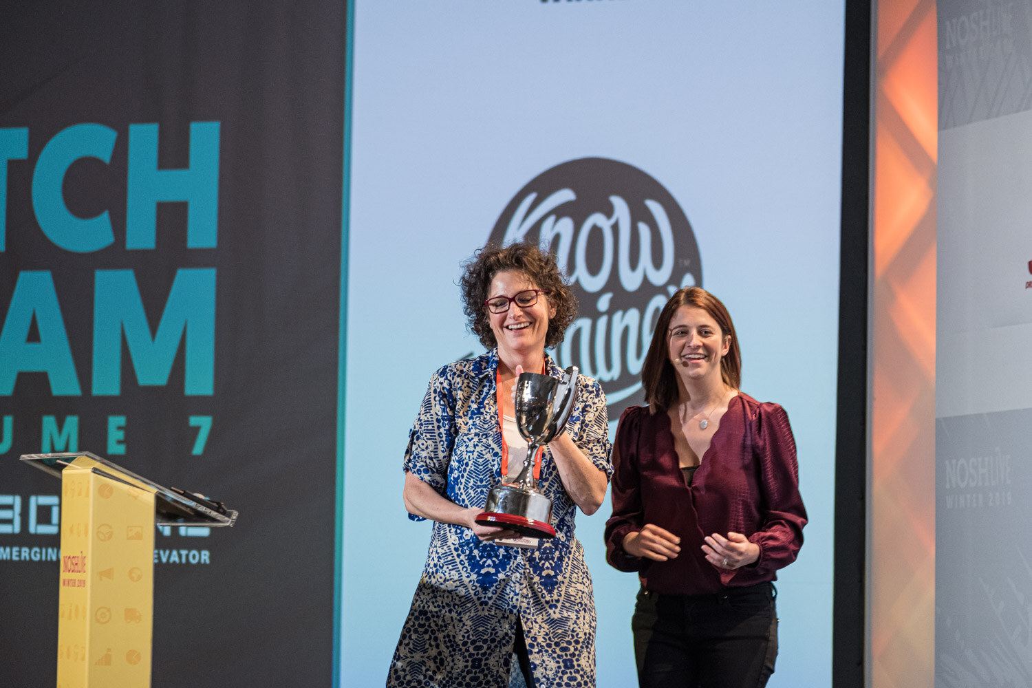 A woman holding a silver trophy beside a host in front of a Pitch Slam Volume 7 screen
