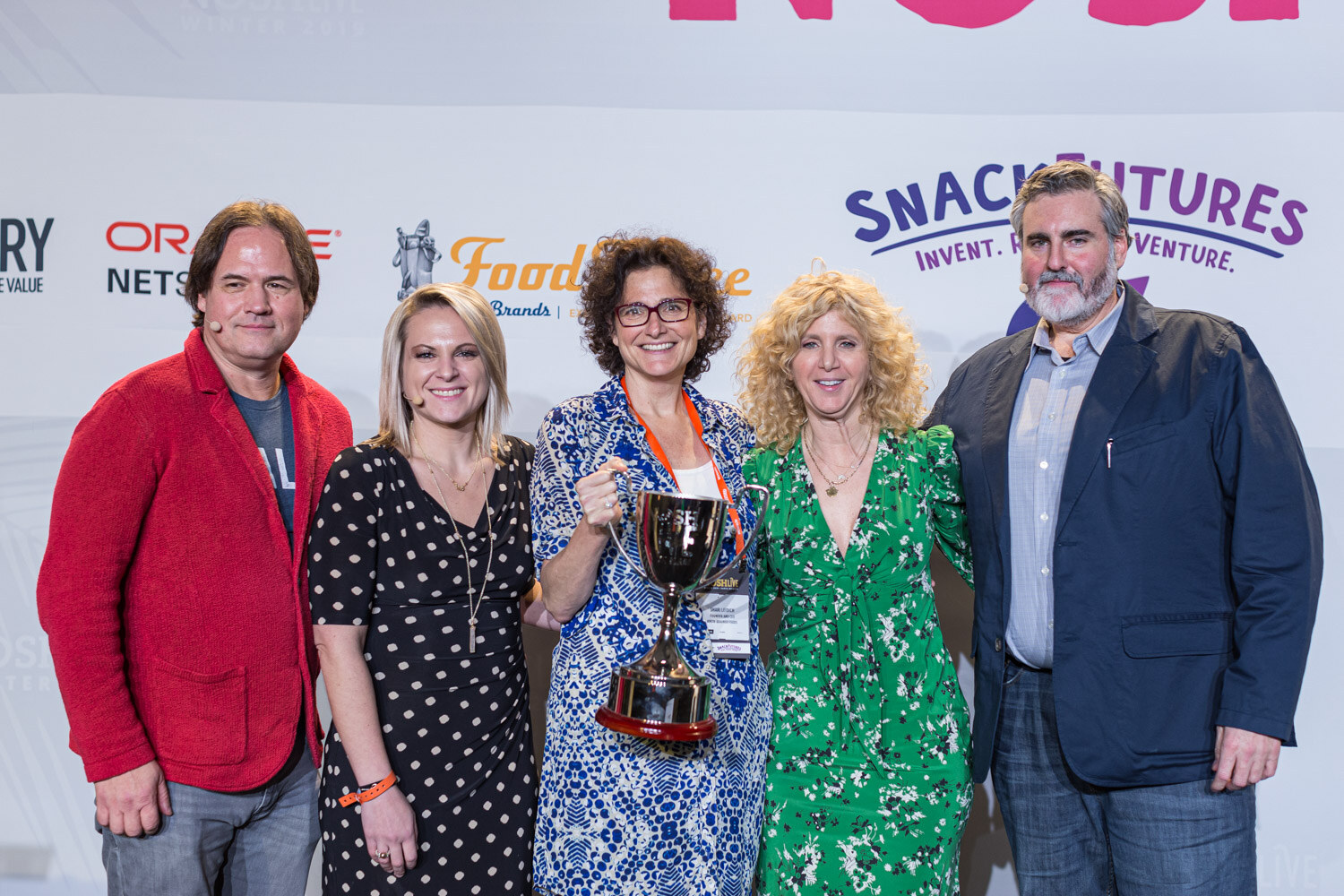 Five people standing together on stage, one holding a silver trophy, in front of the sponsor wall