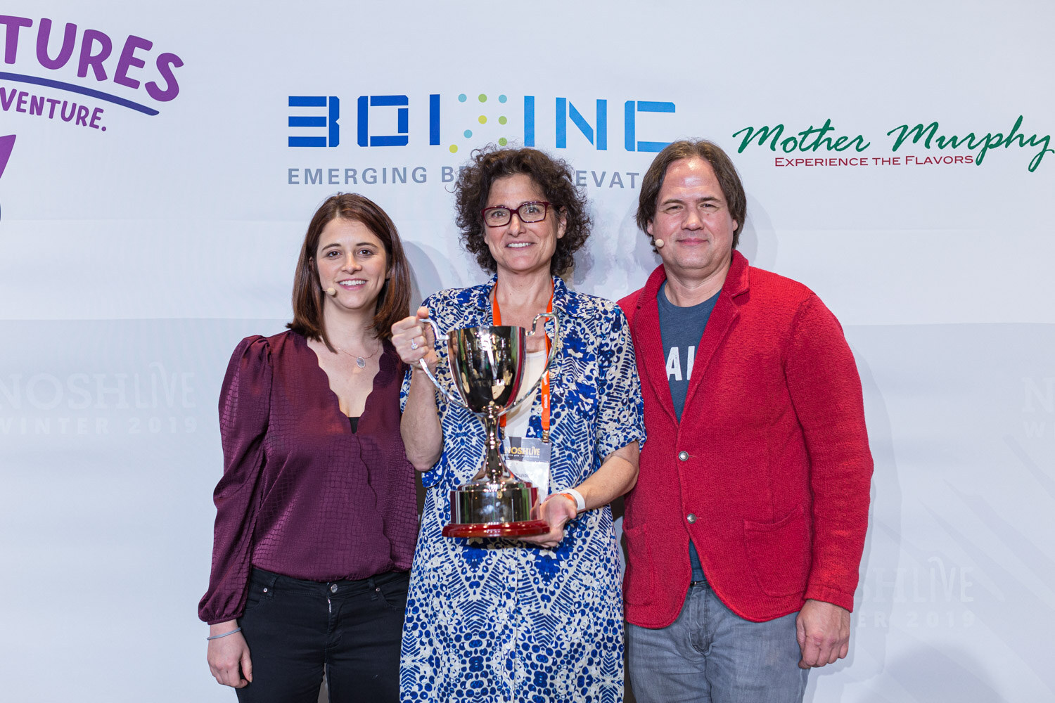 Three people pose on stage with a woman in the middle holding a silver trophy
