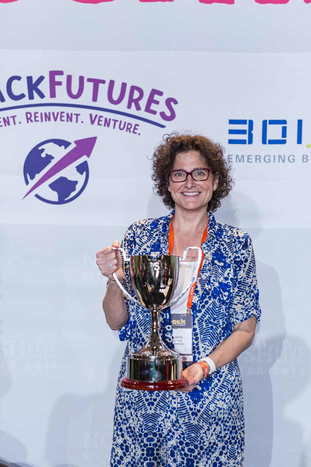 A woman in a blue patterned jacket holding a silver cup in front of a SnackFutures banner