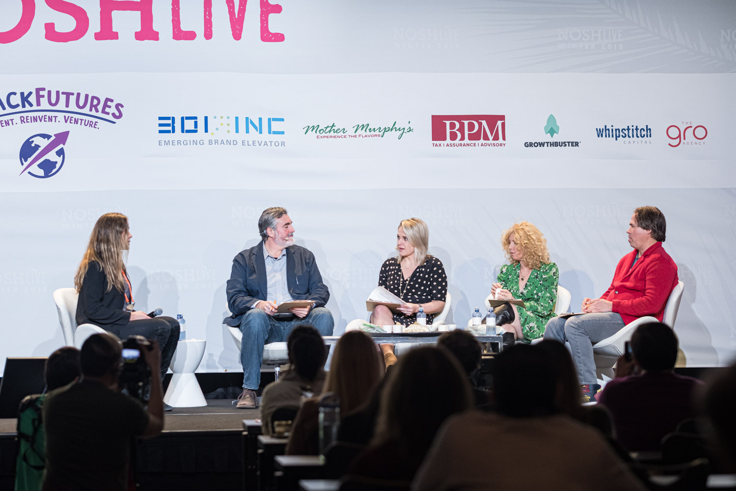 Four judges seated on stage with a presenter on the left and the audience in the foreground