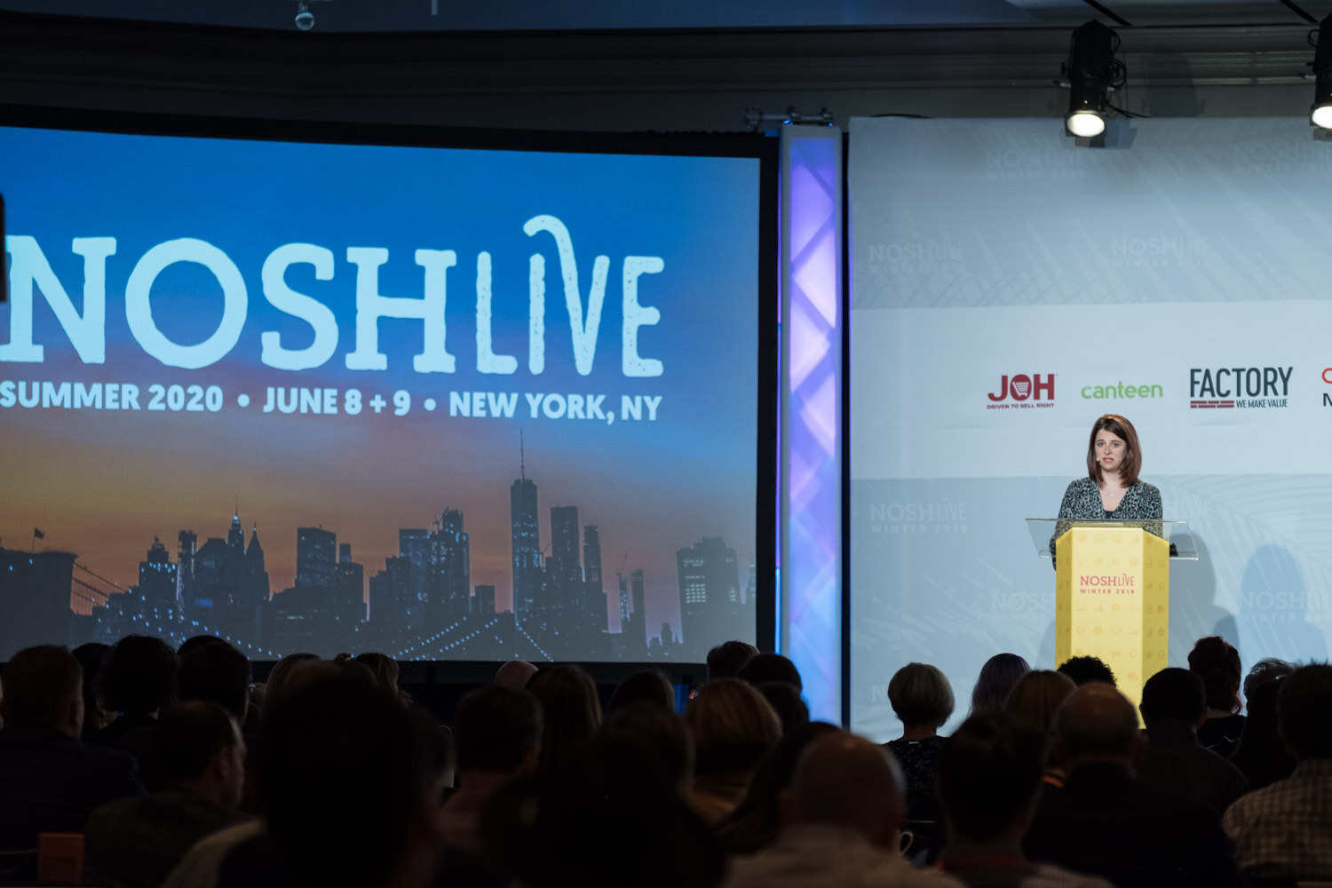 A woman speaks at a yellow lectern on the Nosh Live stage in front of a large screen and a seated audience