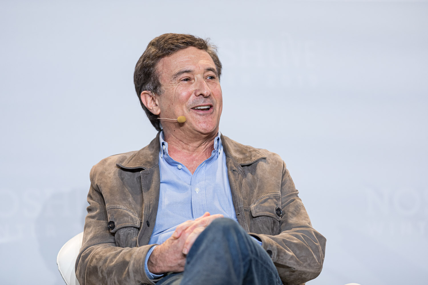 A smiling man in a brown jacket and light blue shirt seated on stage