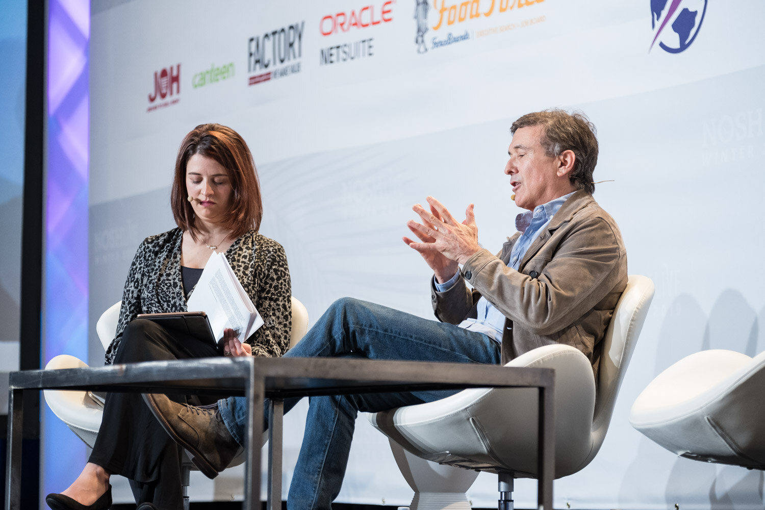 A moderator with a tablet and a man in a brown jacket seated in white chairs on stage