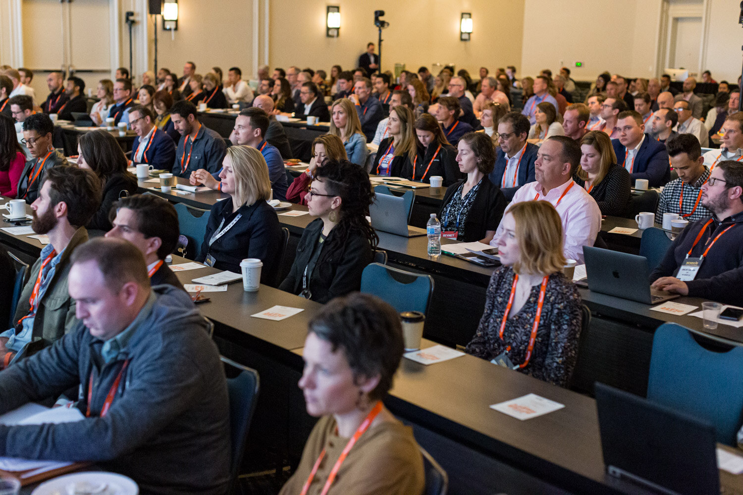 Rows of attendees with orange lanyards seated at long tables in a ballroom