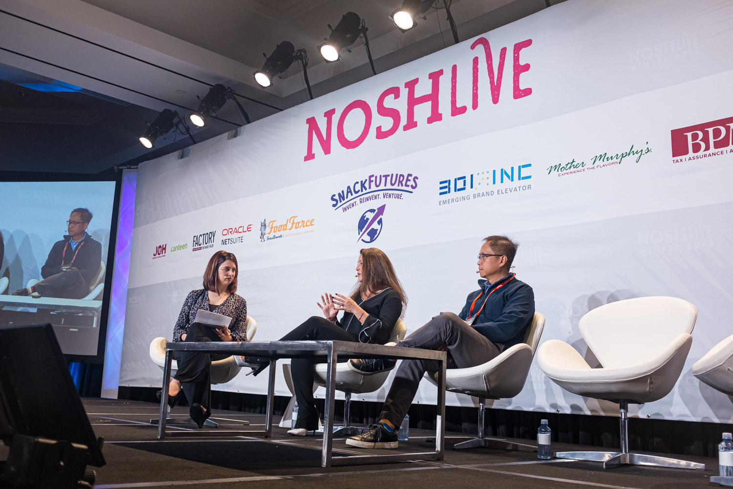 A moderator and two Mondelez presenters seated in white chairs on stage as a screen shows the conversation