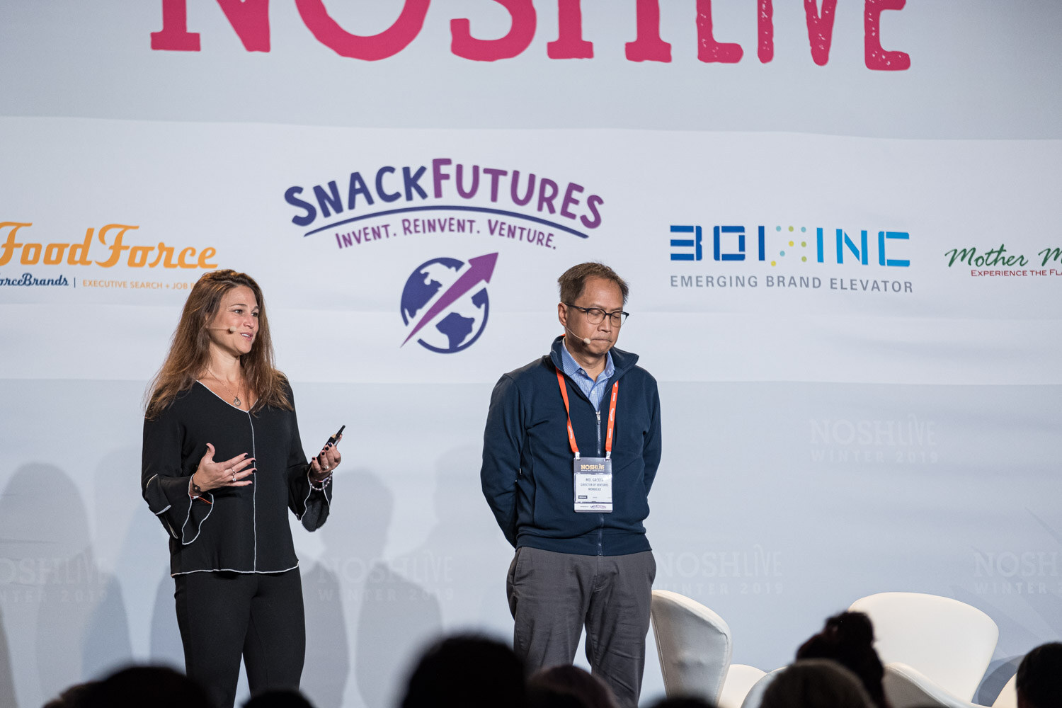 A woman and a man in a navy jacket standing side by side on the Nosh Live stage in front of sponsor logos