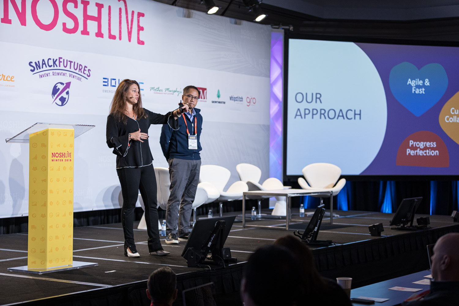 A woman and a man standing at a yellow lectern beside a slide titled Our Approach
