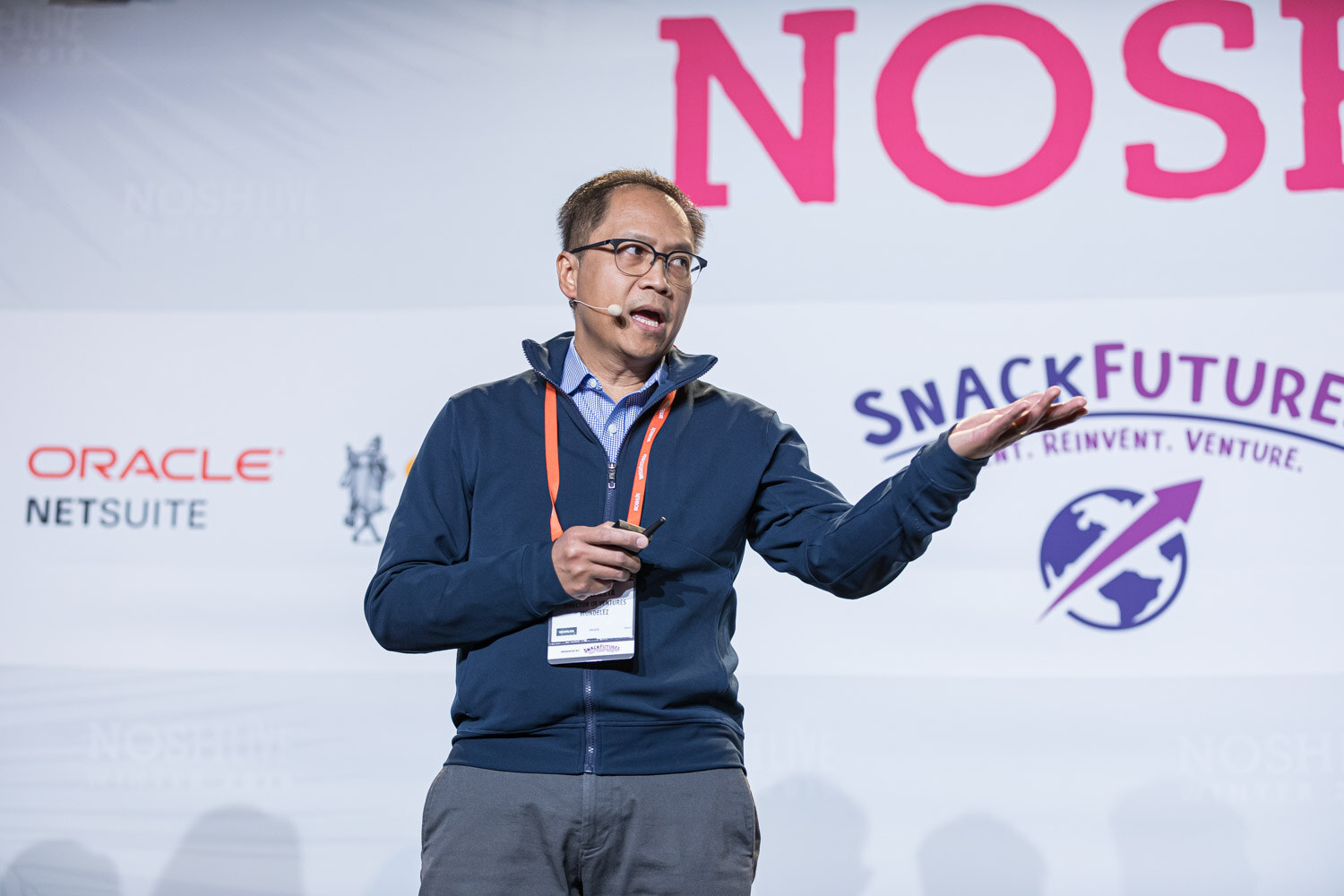 A man with glasses and a headset microphone pointing as he speaks in front of a SnackFutures banner