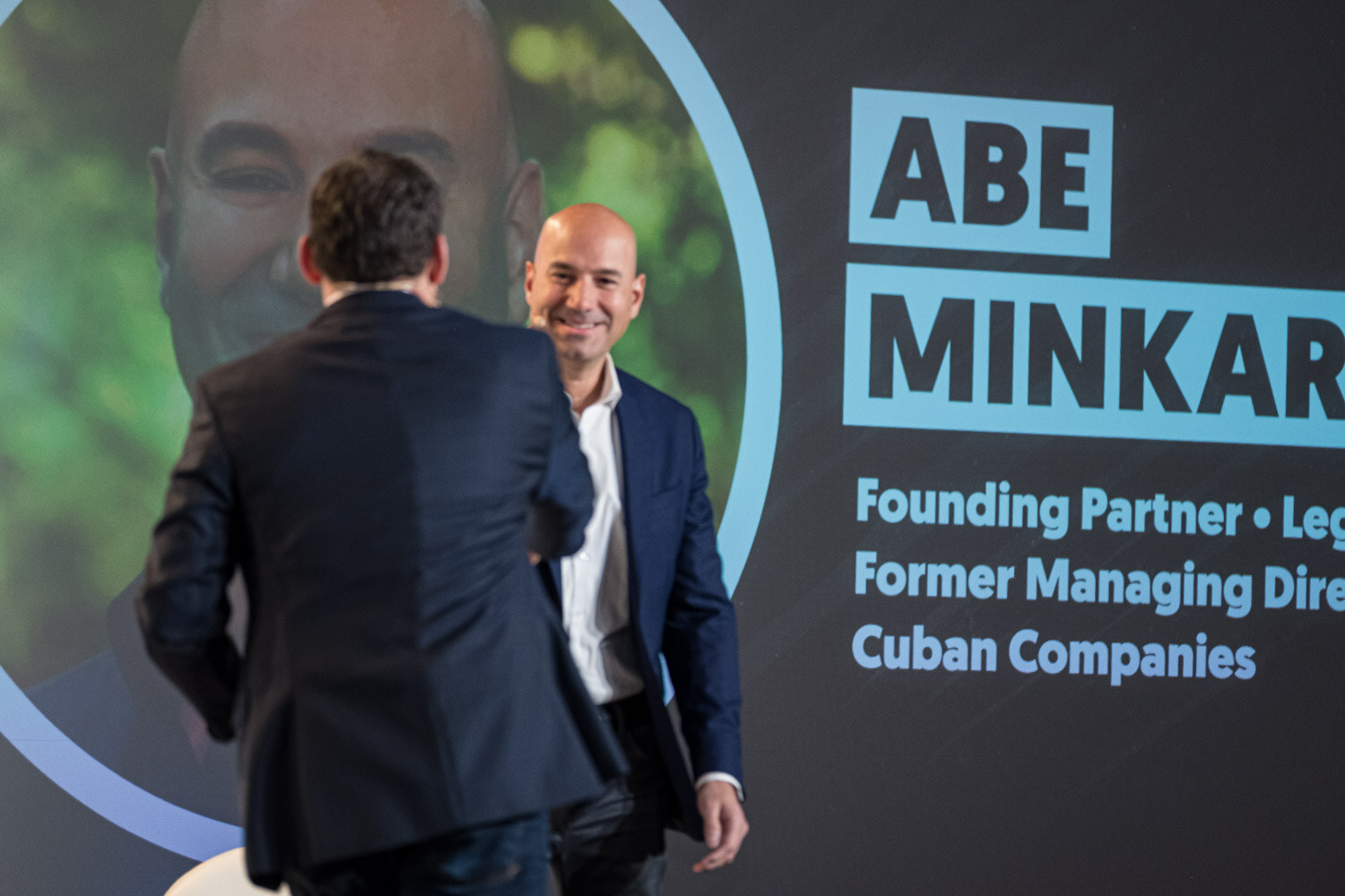 A man in a navy blazer on stage in front of a screen with a title slide reading Abe Minkara, Founding Partner