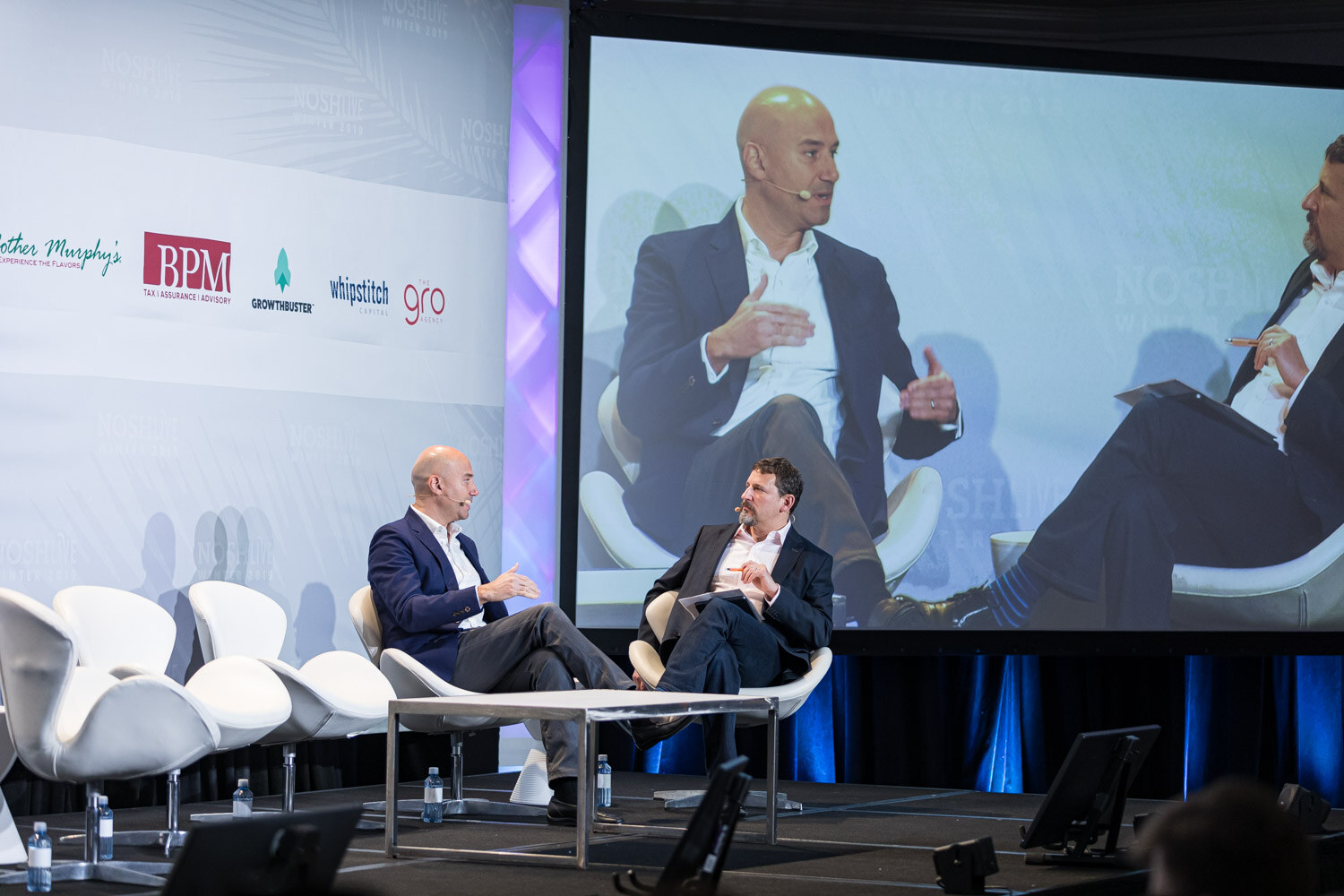 A wide shot of two men on stage with a large screen showing a close-up of the conversation