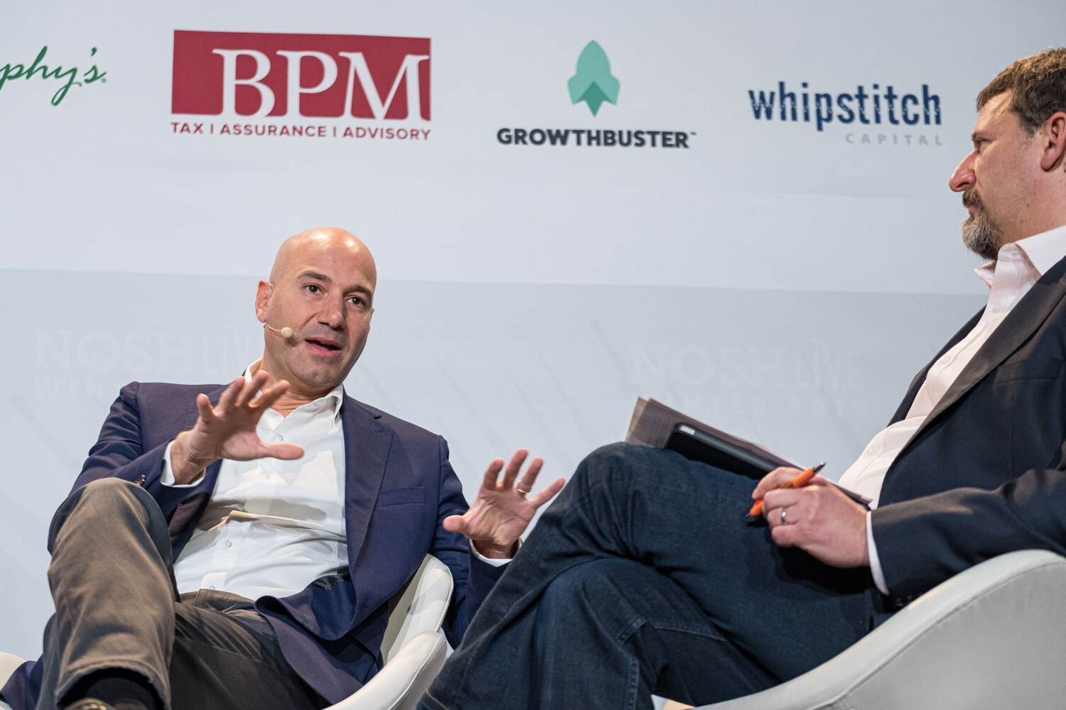 Two men in armchairs on stage, one gesturing while the other holds a tablet, in front of sponsor logos including BPM and Whipstitch Capital