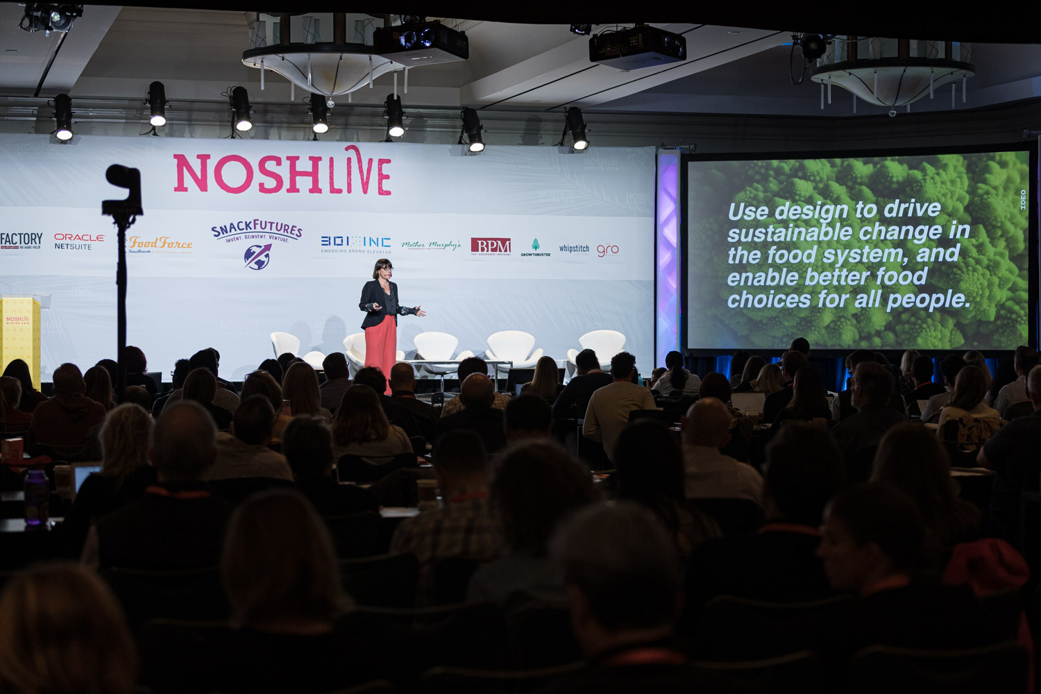 A speaker on stage with a screen reading Use design to drive sustainable change in the food system, and an audience in silhouette