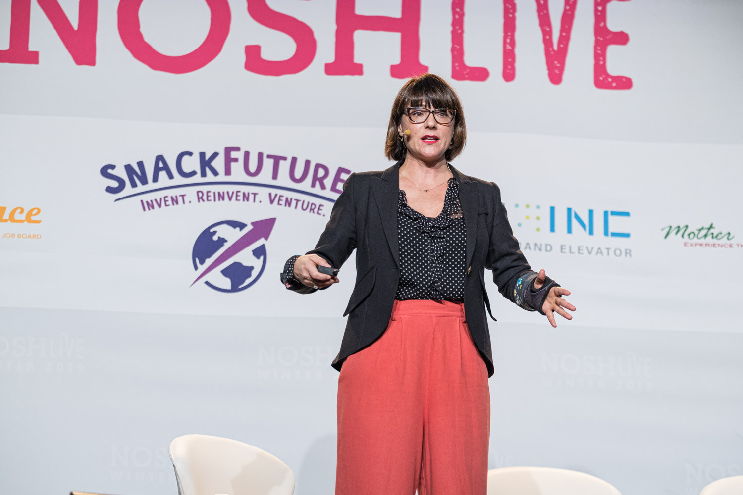 A woman in a black blazer and coral pants gesturing with open arms in front of a SnackFutures banner