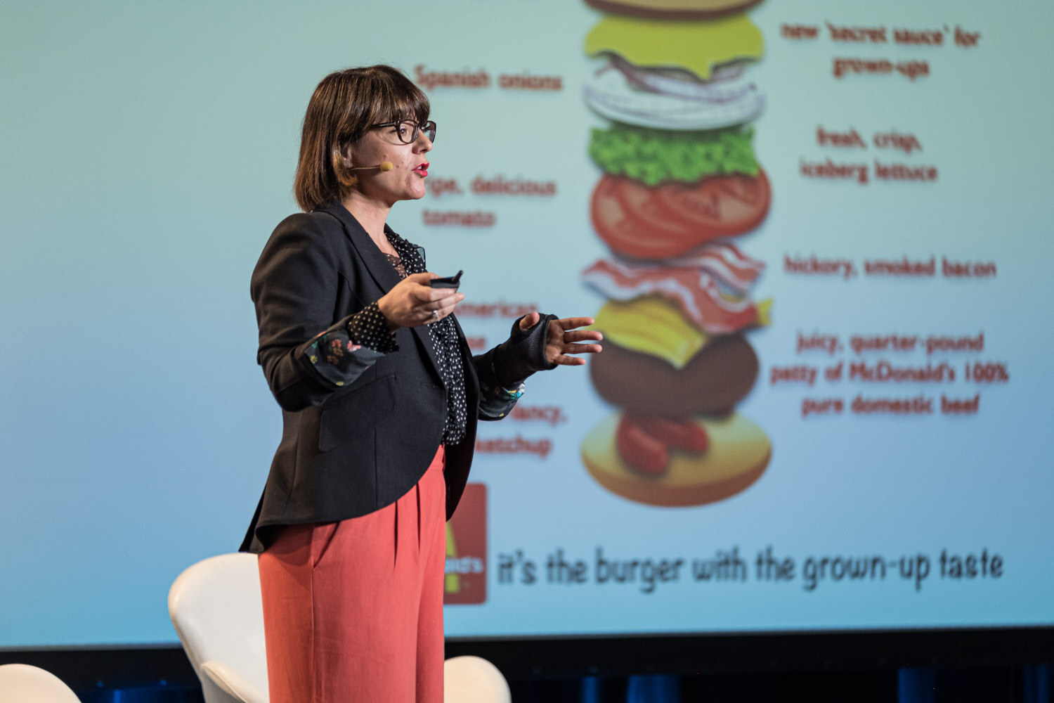 A woman presenting beside a slide showing an illustrated burger layered with ingredient notes