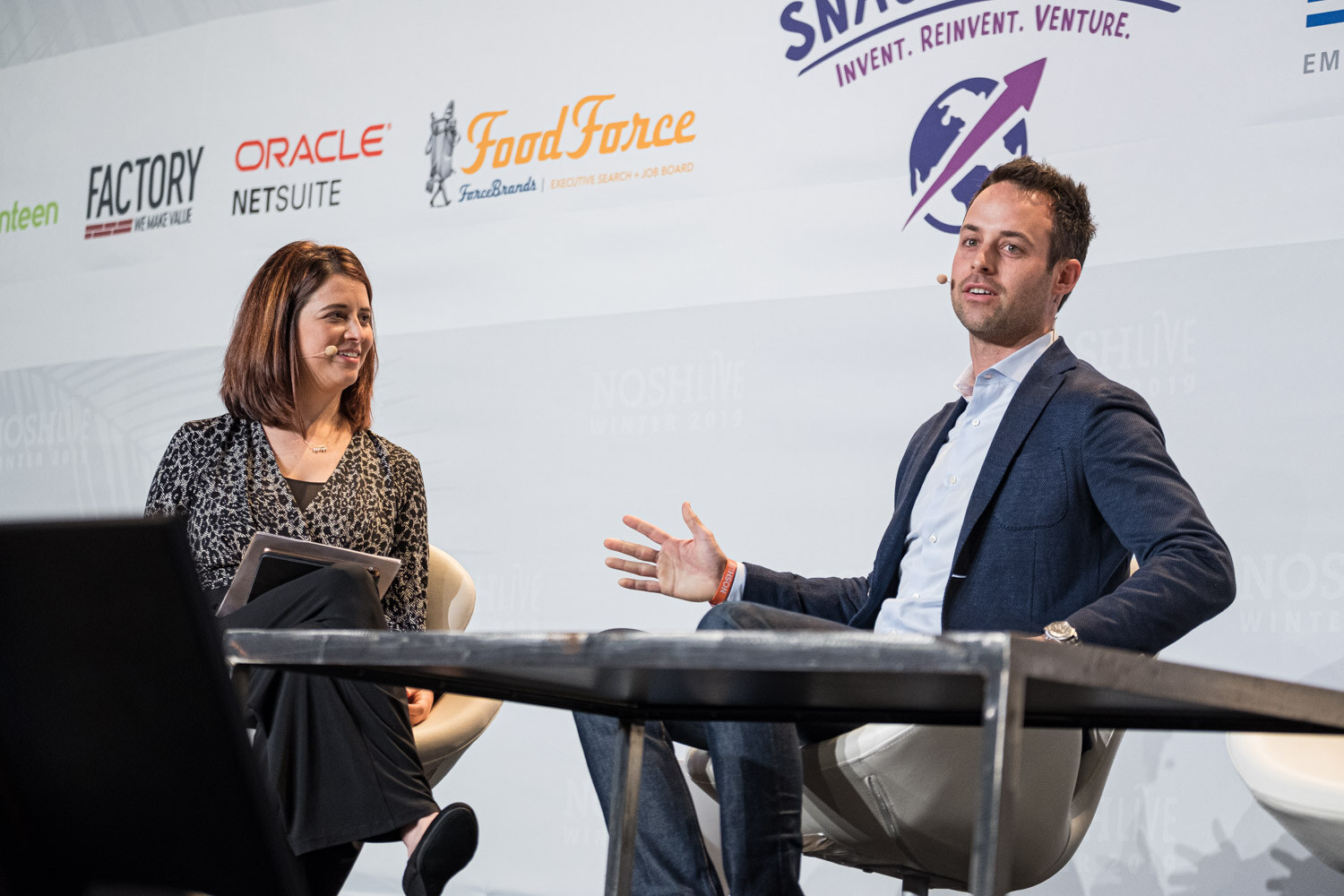 A woman and a man in a navy blazer talking on stage in front of Oracle NetSuite and ForceBrands logos