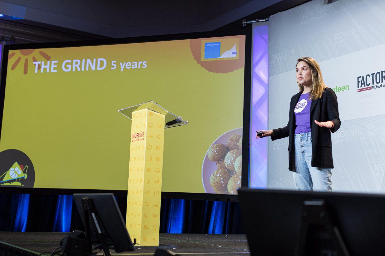 A woman in a purple shirt on stage beside a yellow screen titled The Grind 5 years