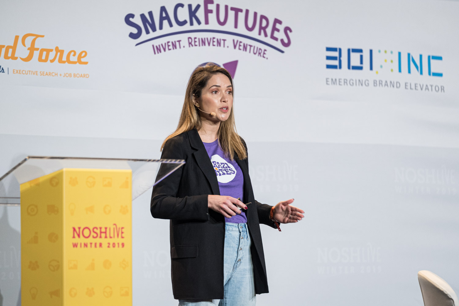 A woman in a purple Brazi Bites shirt and black blazer speaking beside a yellow lectern