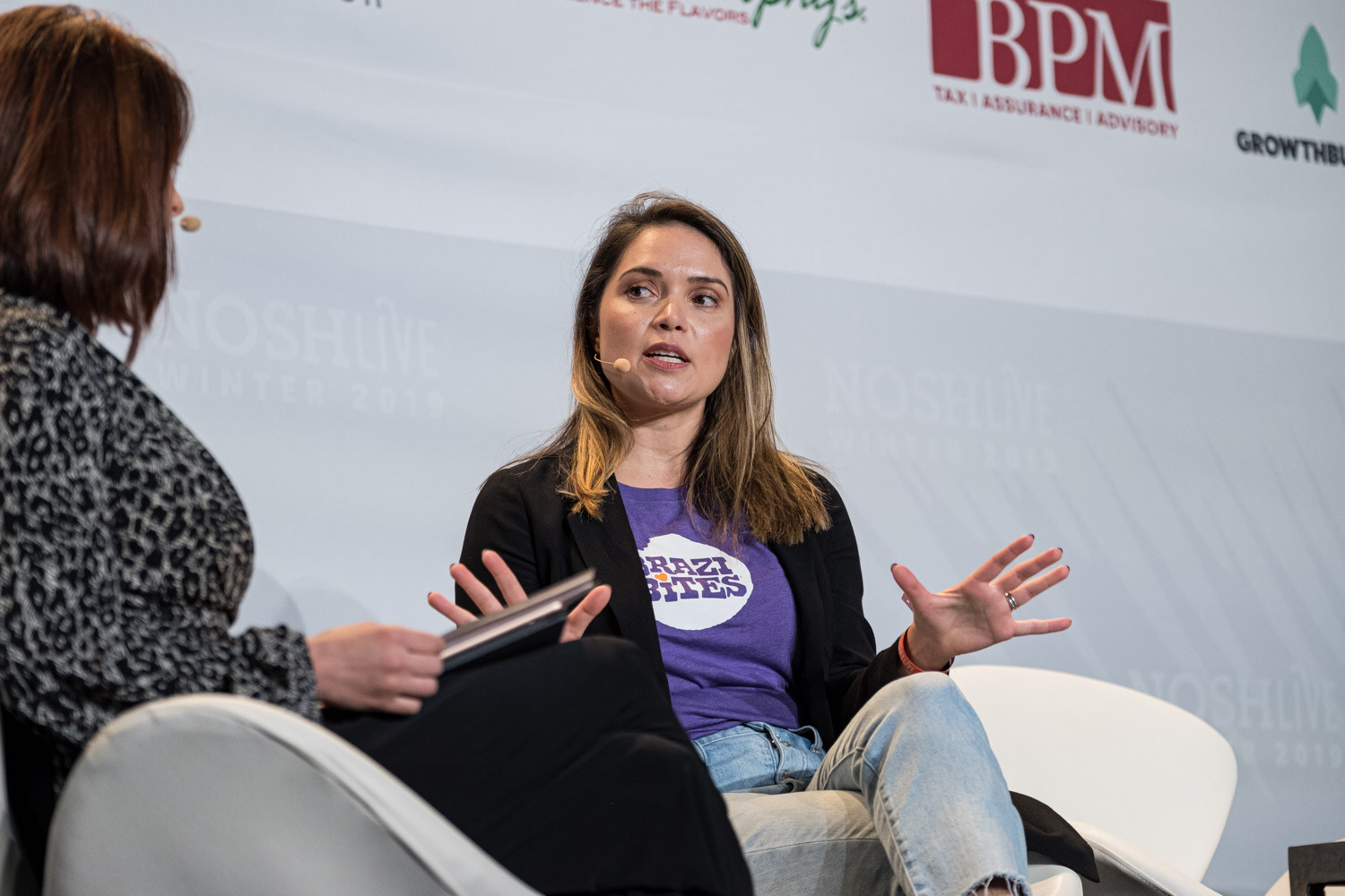 A woman in a purple shirt gesturing while seated across from a moderator