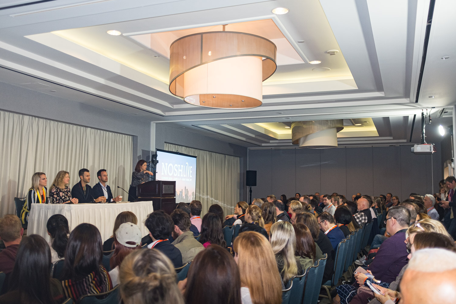 A full audience facing a panel table and a screen in the Loews meeting room
