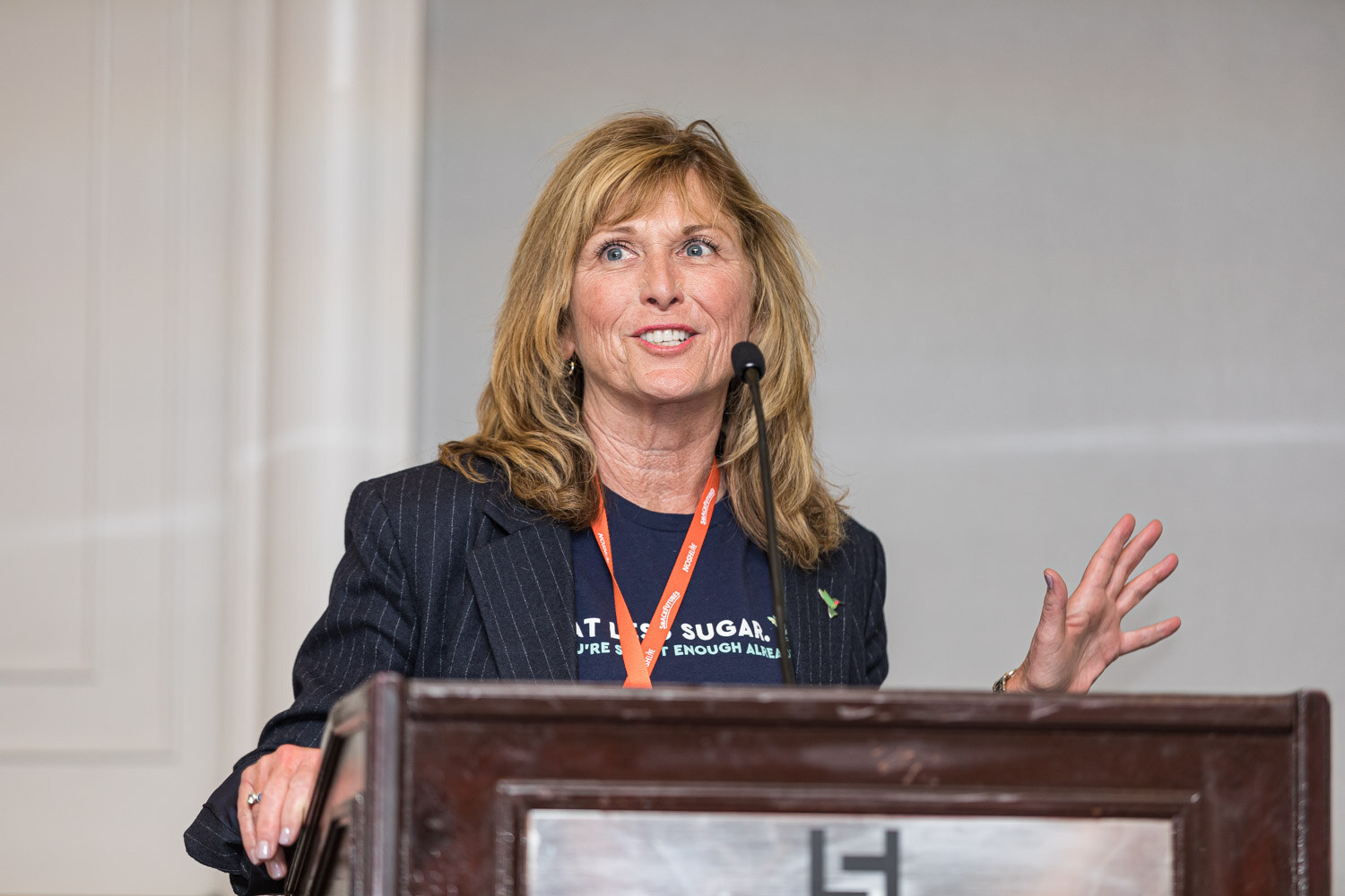 A woman in a navy blazer and Less Sugar T-shirt with an orange lanyard speaking at a lectern