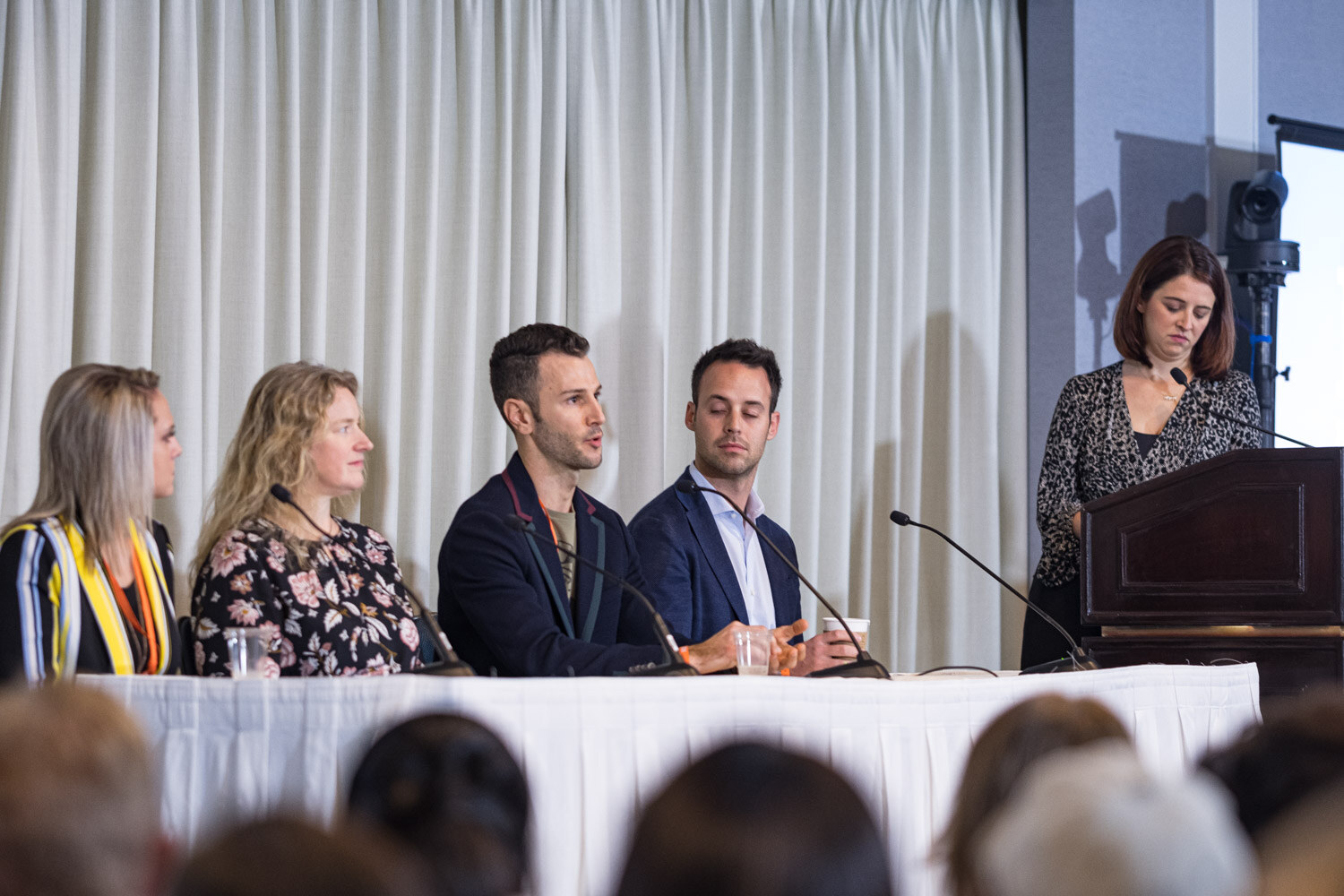 Four panelists seated at a draped table with a moderator standing at a lectern in a hotel meeting room