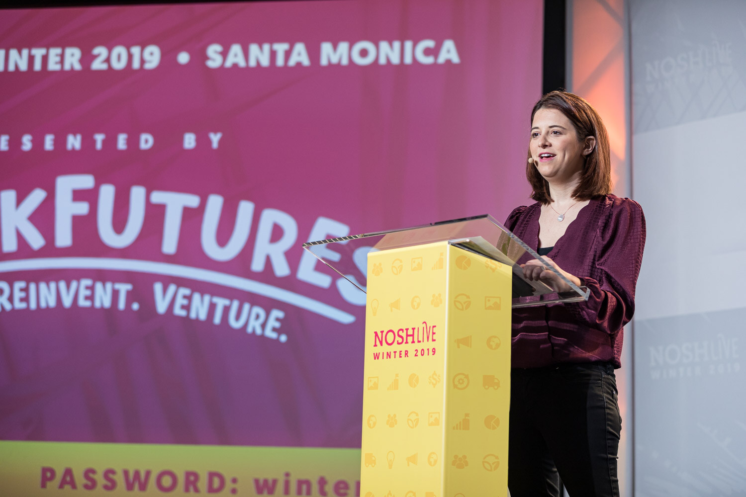 A woman in a maroon top at a yellow lectern in front of a pink Winter 2019 Santa Monica backdrop
