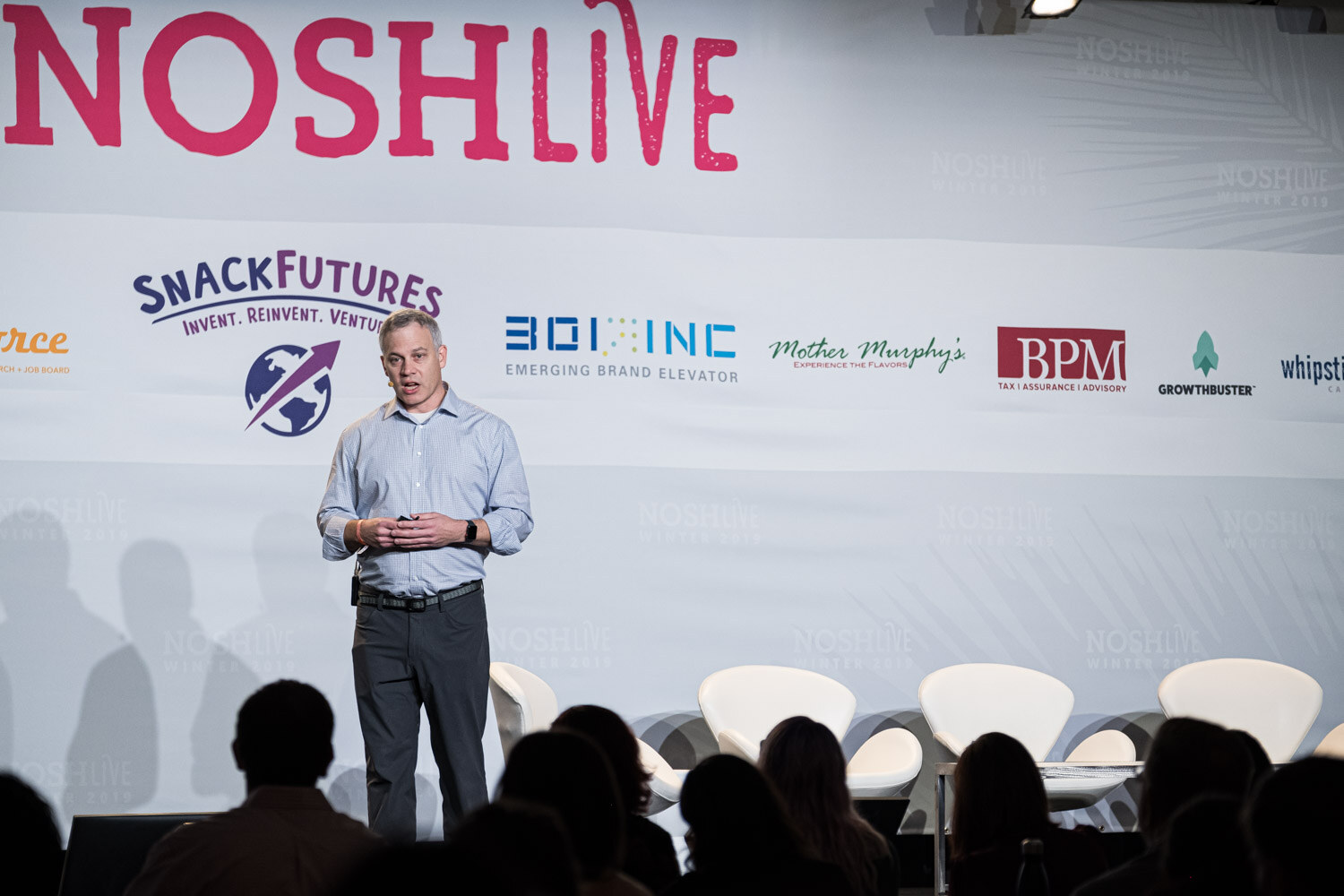 A speaker standing center stage in front of the Nosh Live and sponsor wall with the audience in silhouette
