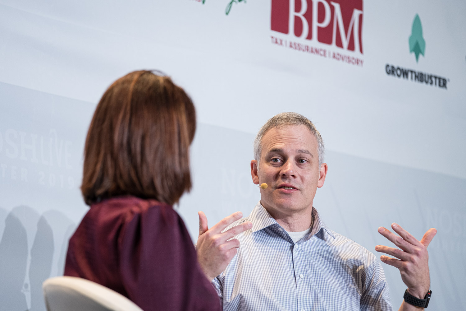 A man gesturing with both hands while a moderator in maroon listens beside him