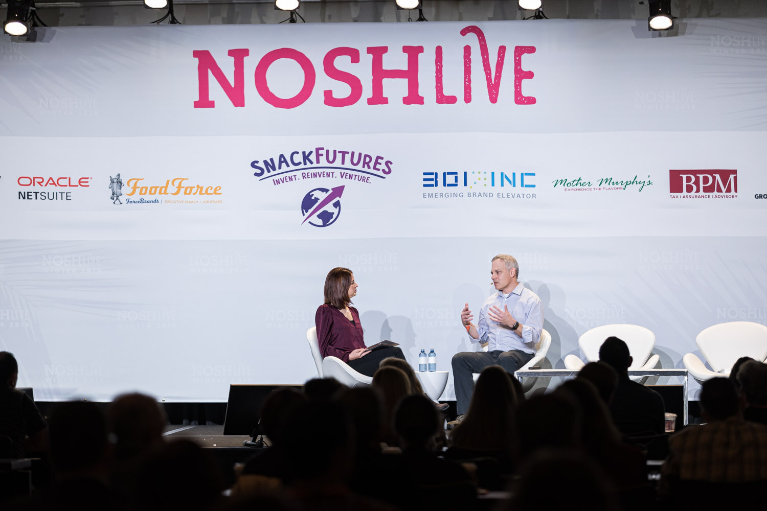 Two people in chairs on stage under the Nosh Live and sponsor banner with the audience in silhouette