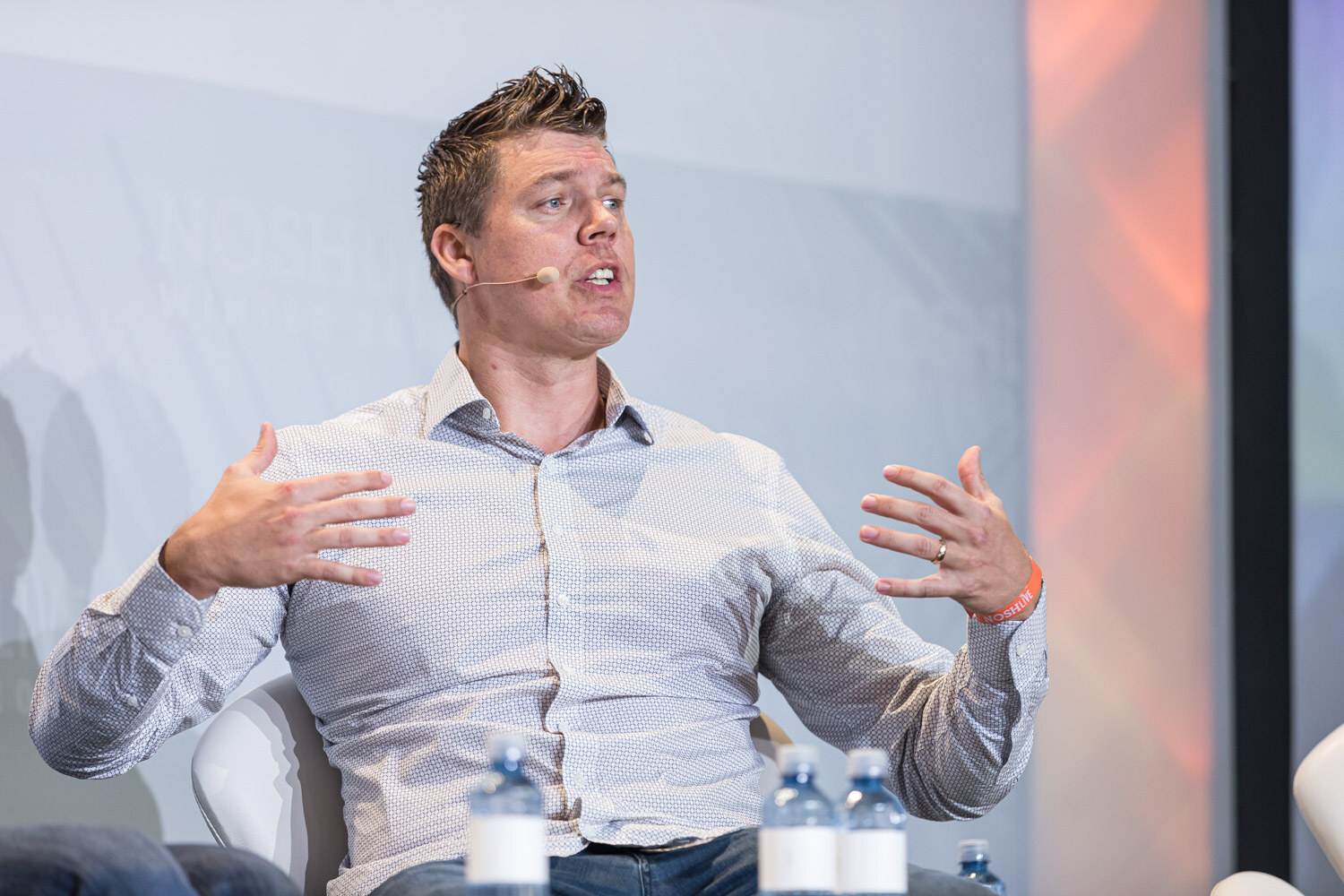 A man in a light patterned shirt with a headset microphone gesturing while seated on stage