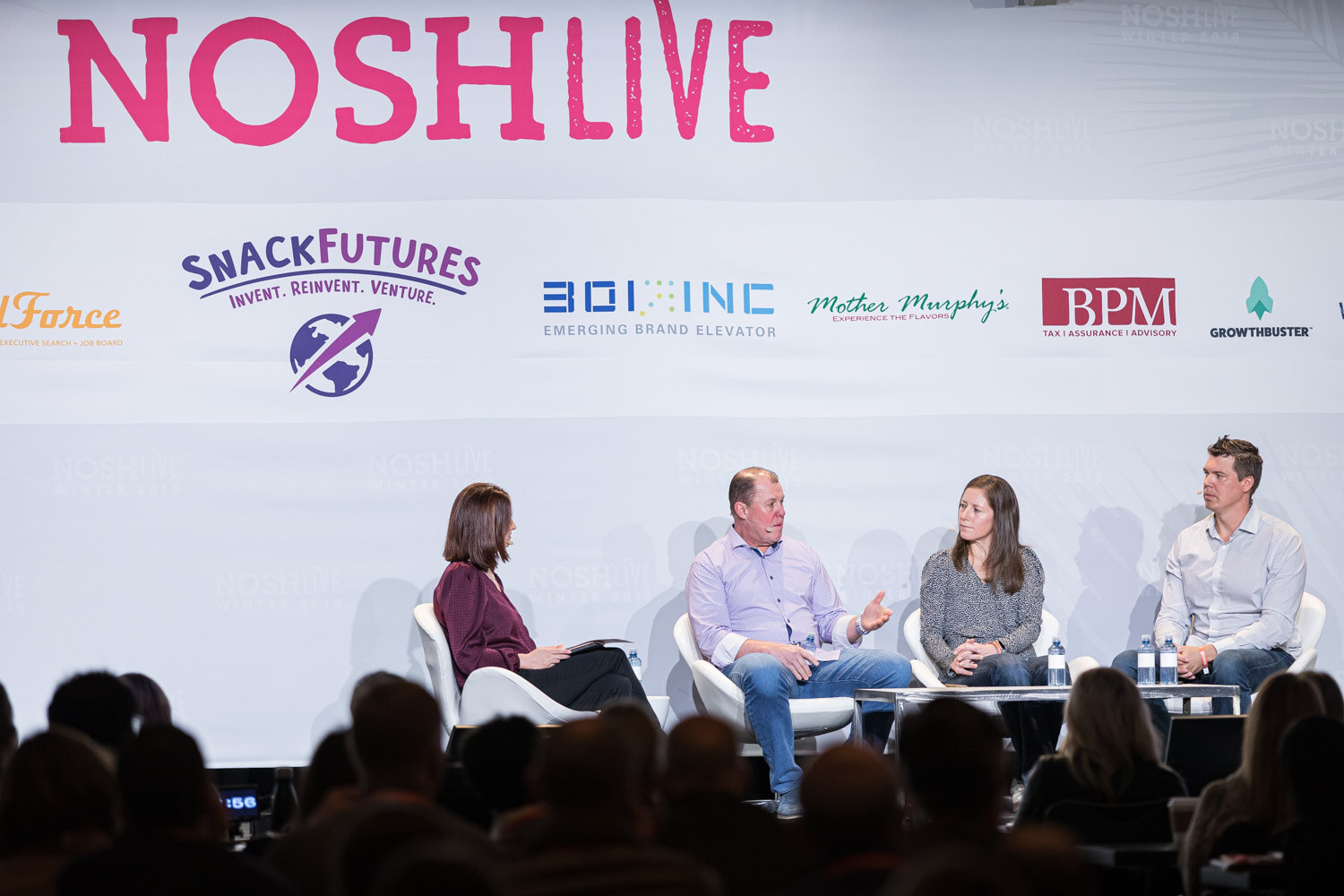 Four people seated on stage under the Nosh Live banner with silhouettes of the audience in front