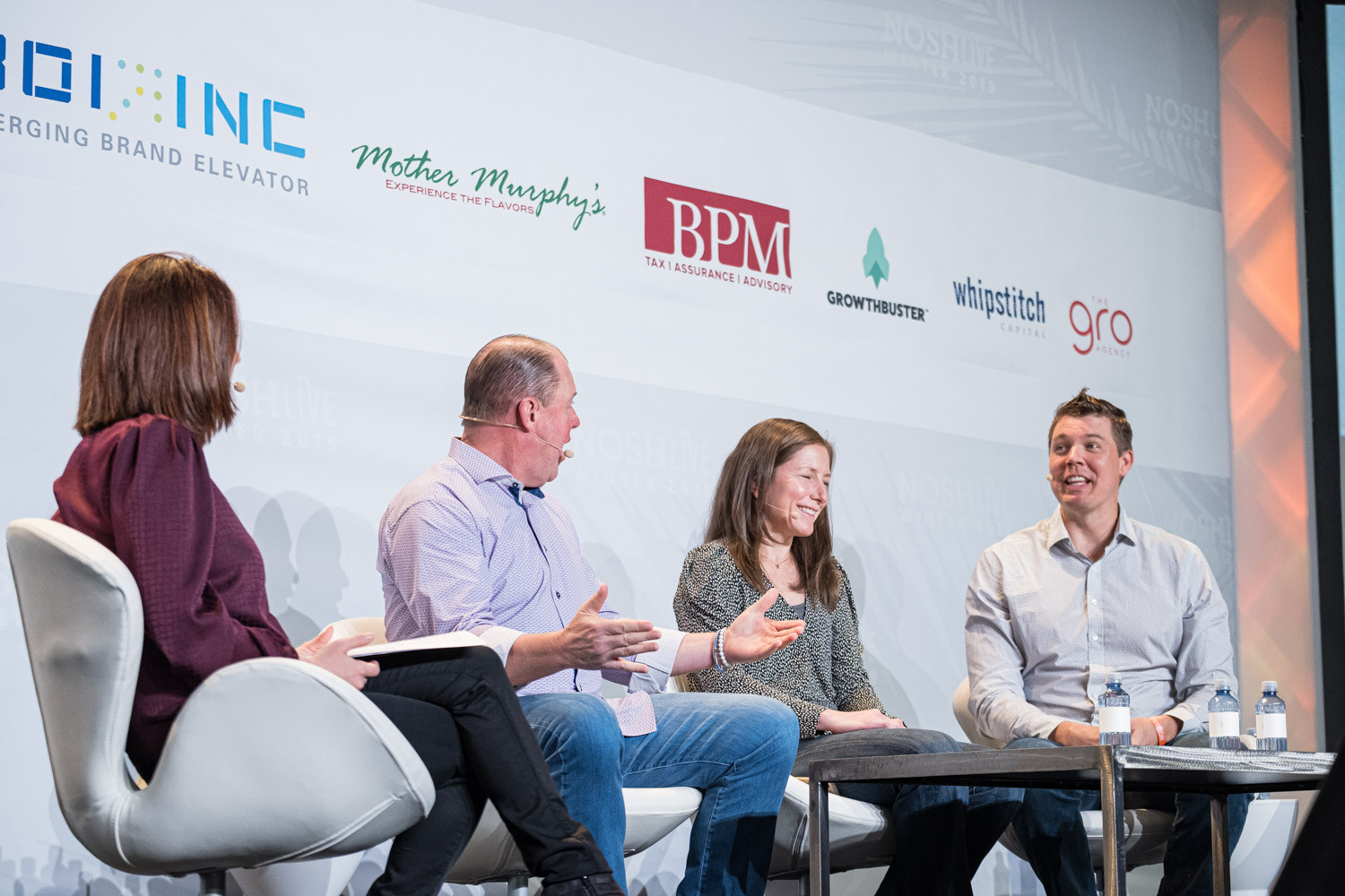 A moderator in maroon facing three panelists seated around a low table on stage