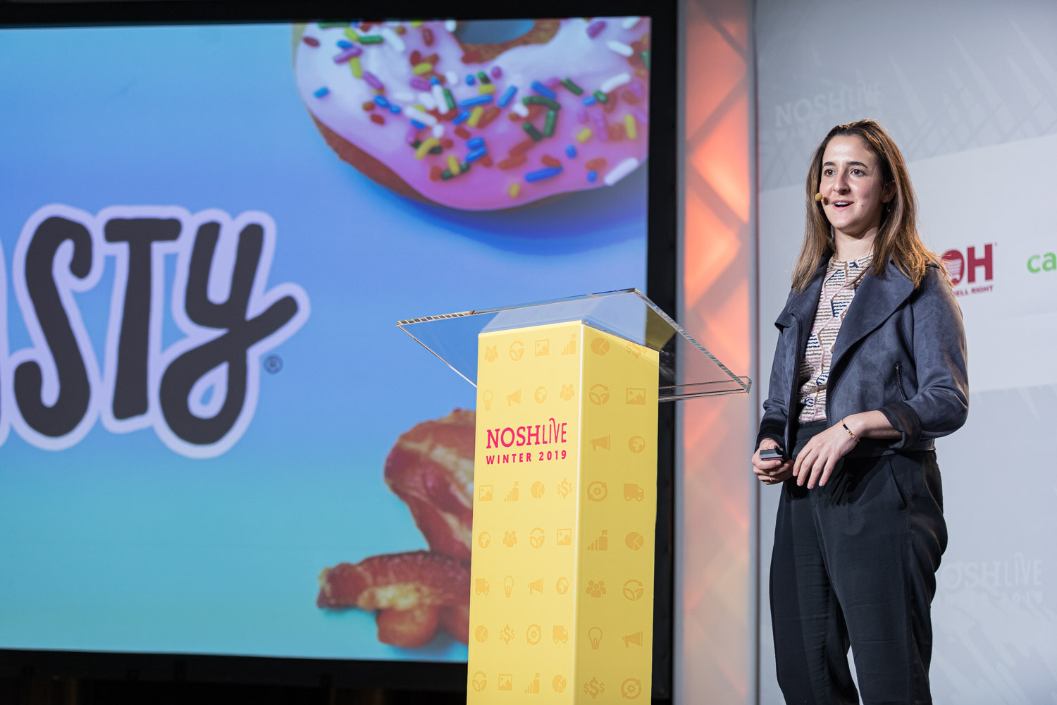 A woman standing beside a yellow lectern next to a screen showing a sprinkled donut