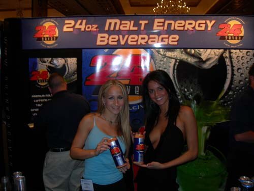 Two women hold cans of 24 Seven malt energy beverage in front of a booth banner reading 24oz. Malt Energy Beverage, with a man standing behind them.